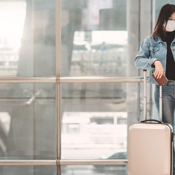 Young woman wearing face mask uses her phone while standing with luggage in airport