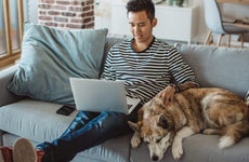 Young man at home uses his laptop on couch with his dog
