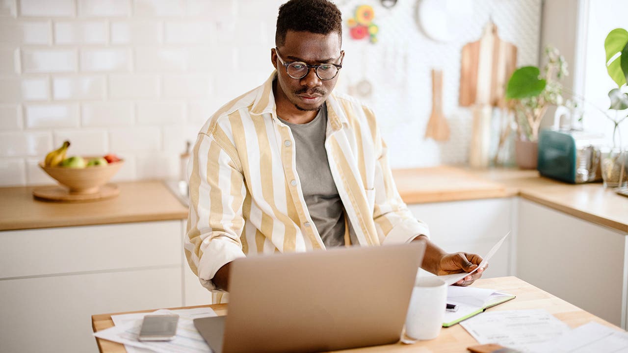 African man working from home, using laptop, paying utility bills. Young man doing taxes at home. Man talking to costumer service.