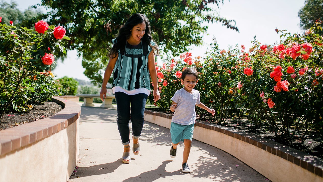 A mother and child run along a pathway with flowers and hedges along the side.