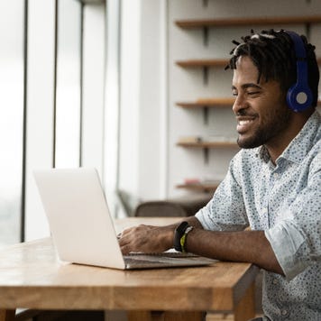 A smiling young man sits at a desk with his laptop