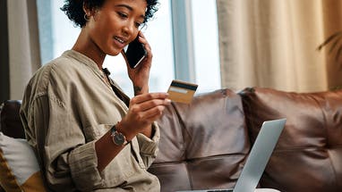 Shot of a young woman looking at her credit card while talking on the phone and sitting with her laptop