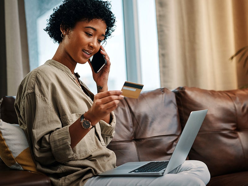 Shot of a young woman looking at her credit card while talking on the phone and sitting with her laptop