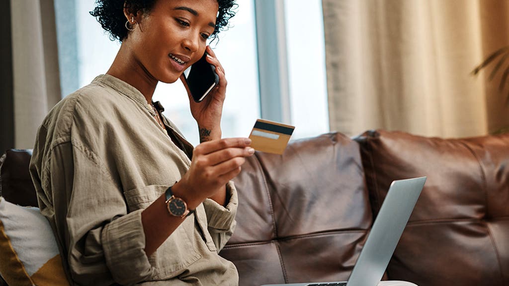 Shot of a young woman looking at her credit card while talking on the phone and sitting with her laptop