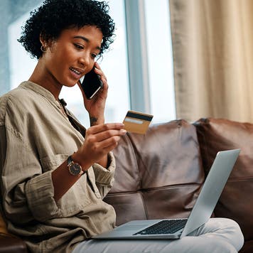 Shot of a young woman looking at her credit card while talking on the phone and sitting with her laptop