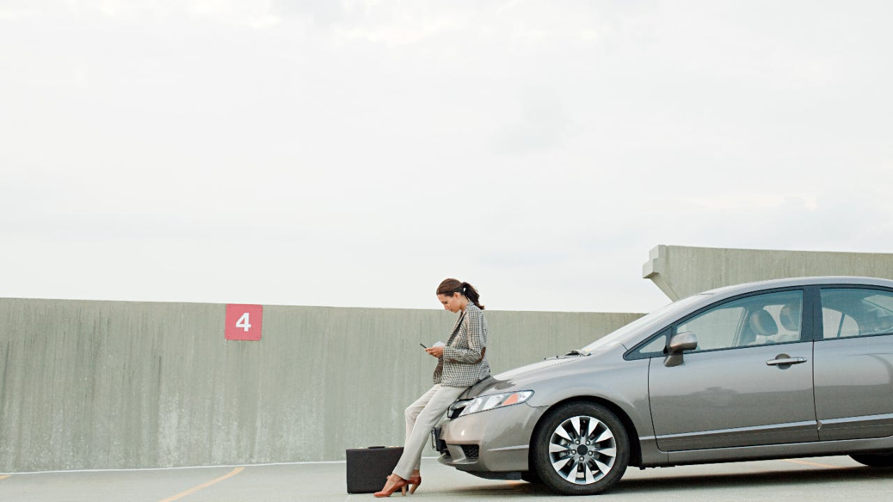 Business woman texting while leaning on the hood of her silver sedan in a car park