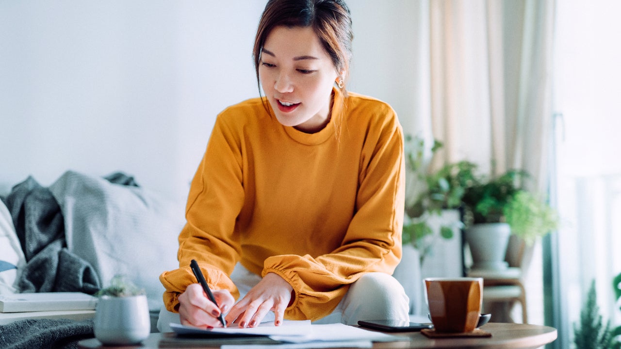 Young Asian woman holding a pen and signing paperwork in the living room at home.
