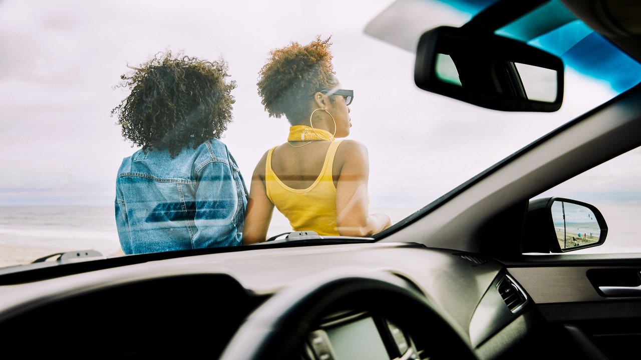 Two black women are leaning against the hood of their car enjoying a nice day.