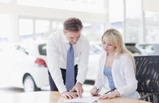 Woman sits at table filling out paperwork with man standing to her left pointing something out, cars are in the background