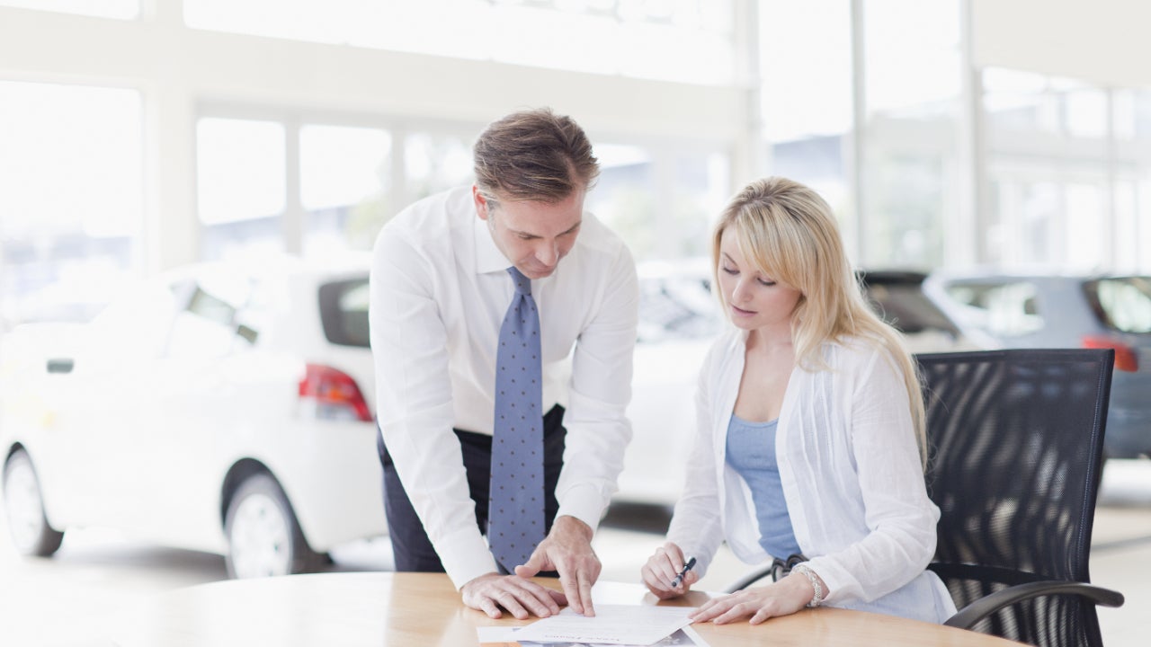 Woman sits at table filling out paperwork with man standing to her left pointing something out, cars are in the background