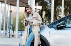 Smiling woman in beige overcoat looking at phone while leaned against silver car
