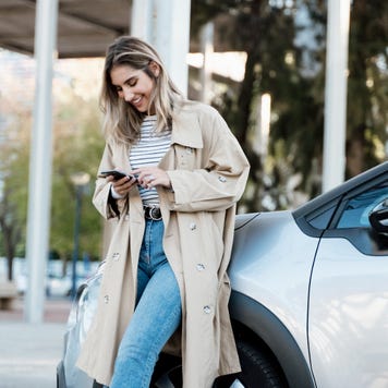 Smiling woman in beige overcoat looking at phone while leaned against silver car