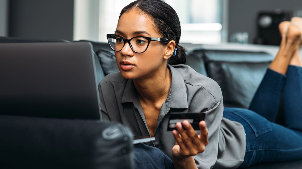 woman laying on couch while working on laptop and holding credit card