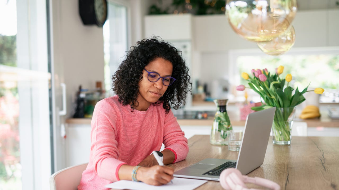 Multiracial woman having home office in kitchen, writing notes.