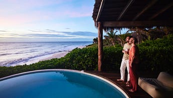couple standing poolside watching sunrise at luxury tropical villa overlooking beach and ocean