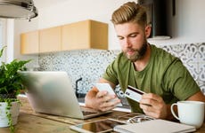 man sitting in kitchen looking at his phone and holding credit card