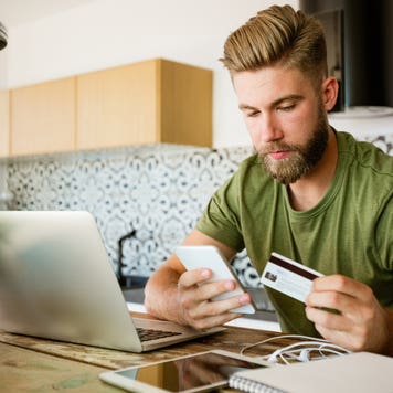 man sitting in kitchen looking at his phone and holding credit card