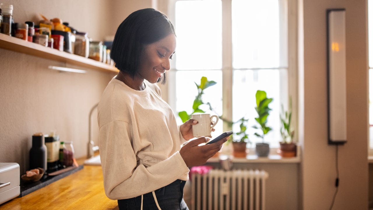 Woman leaning on counter at home, holding a mug, using her phone and smiling