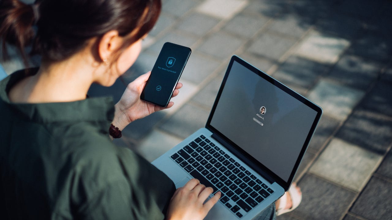 Woman sitting on the bench in an urban park working outdoors, logging in to her laptop and holding smartphone on hand with a security key lock icon on the screen. Privacy protection, internet and mobile security concept