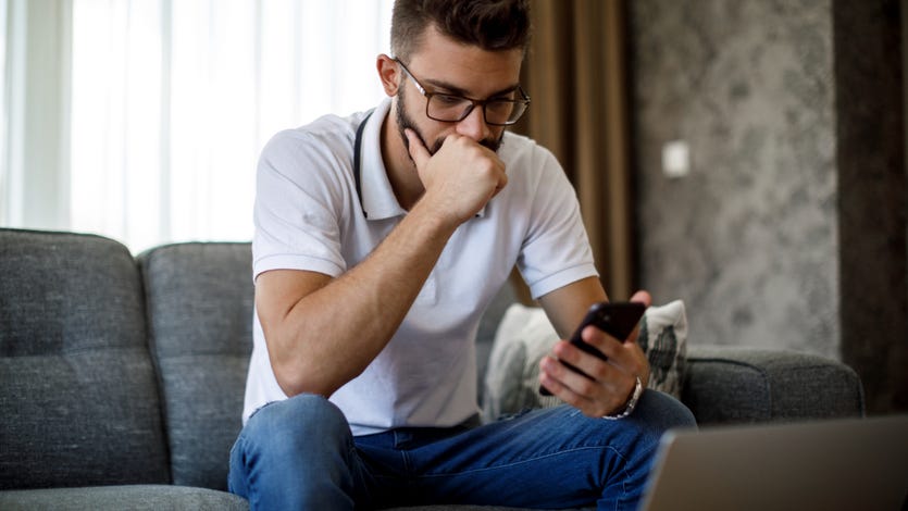 Man with glasses sitting on couch looking pensively at phone