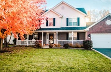 White and brick suburban home with white front porch, pumpkin on stoop and bright orange fall leaves on tree in front yard