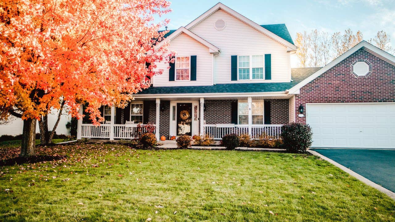 White and brick suburban home with white front porch, pumpkin on stoop and bright orange fall leaves on tree in front yard