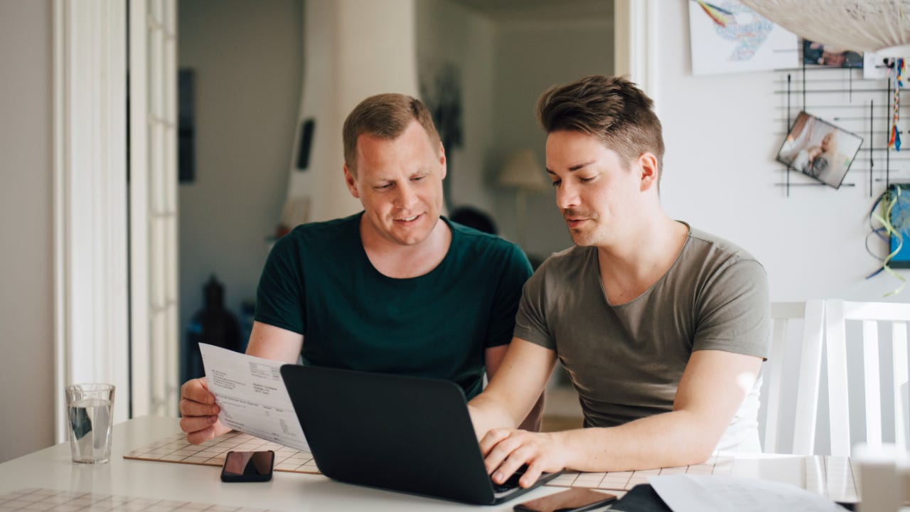 A couple in their home, one man is on a laptop, the other man is holding a paper and smiling