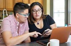 Two people sitting together in family home with paperwork, laptop, and making calculations on smart phone