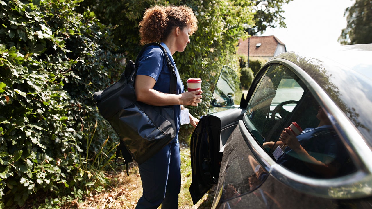 Driver holding a coffee and a satchel getting into their car. From the attire, it would appear this person is a nurse or some other kind of aid worker.