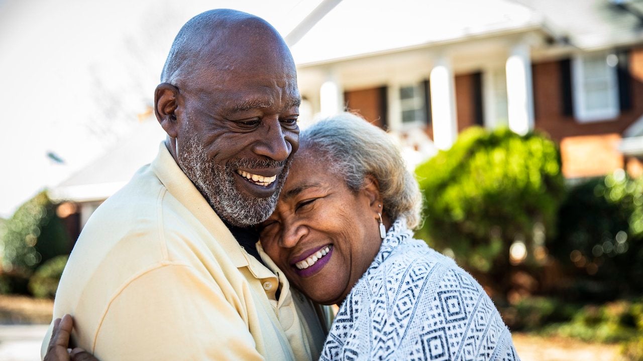 Older african-american couple smiling and embracing in front of a brick home with white pillars