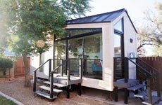 A tiny house with large glass windows, sits in the backyard surrounded by a wooden fence and trees.