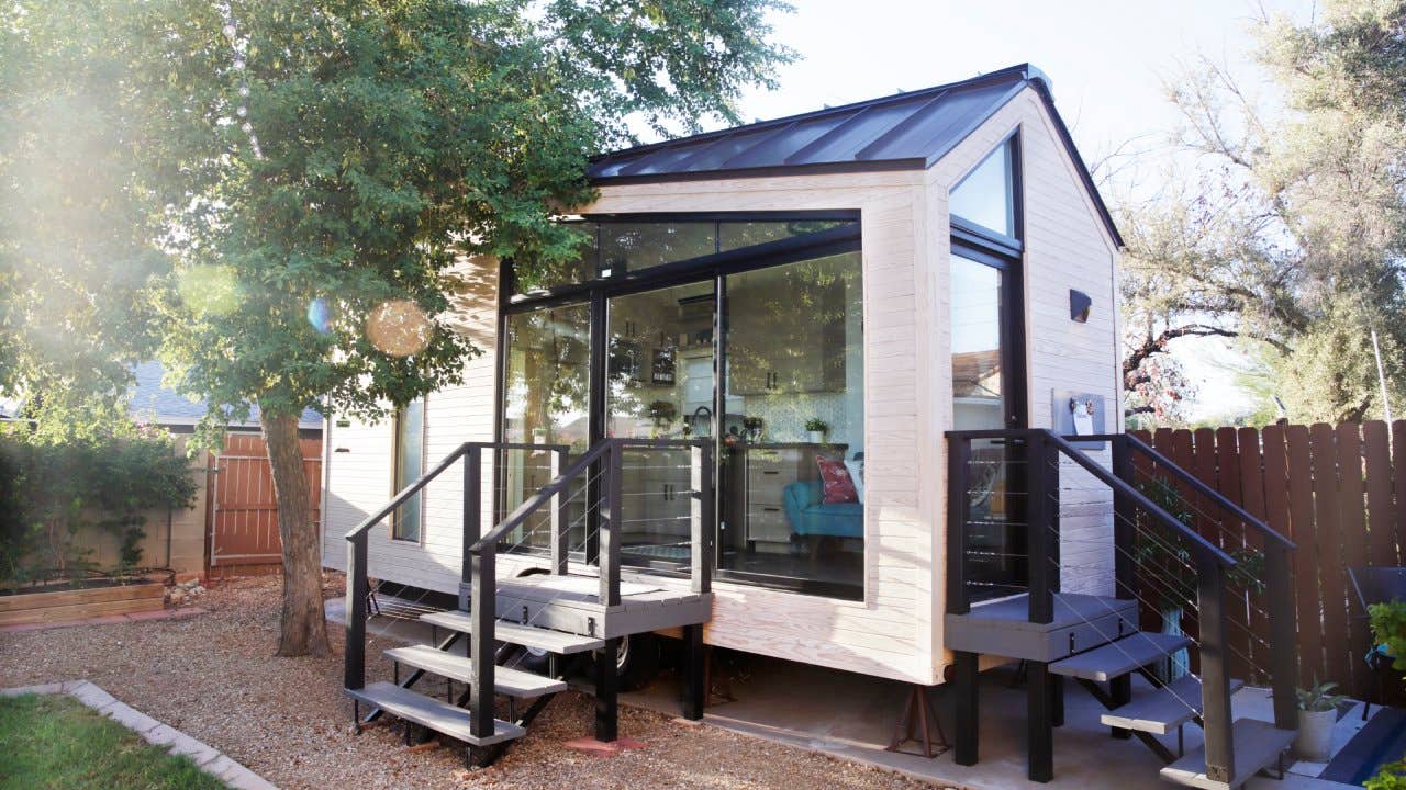 A tiny house with large glass windows, sits in the backyard surrounded by a wooden fence and trees.