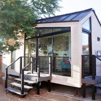 A tiny house with large glass windows, sits in the backyard surrounded by a wooden fence and trees.