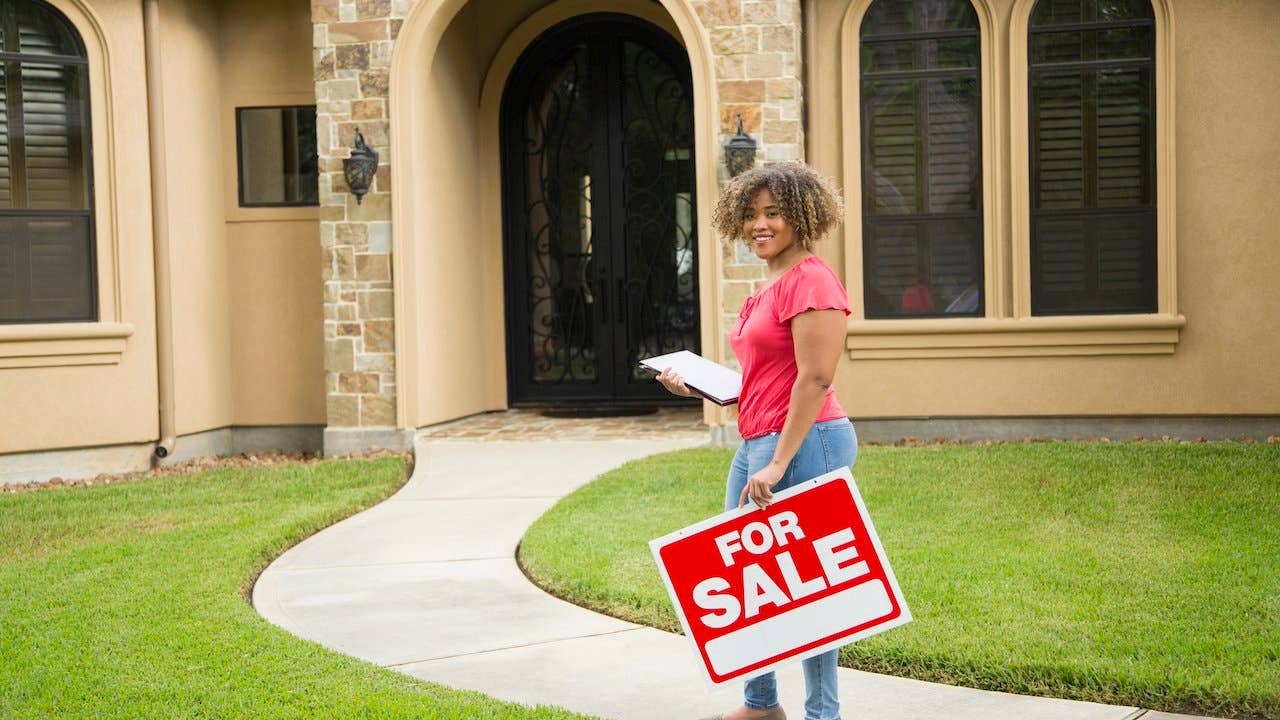 Realtor stands outside suburban home with clipboard and for sale sign