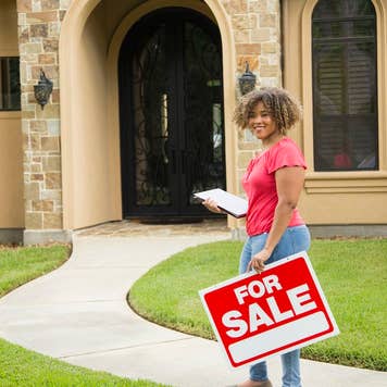 Realtor stands outside suburban home with clipboard and for sale sign