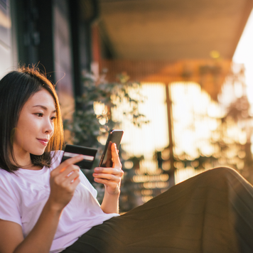 Smiling young Asian woman using smart phone and credit card to mange online banking, sitting on the balcony with dramatic sunlight. Online shopping makes life easier.