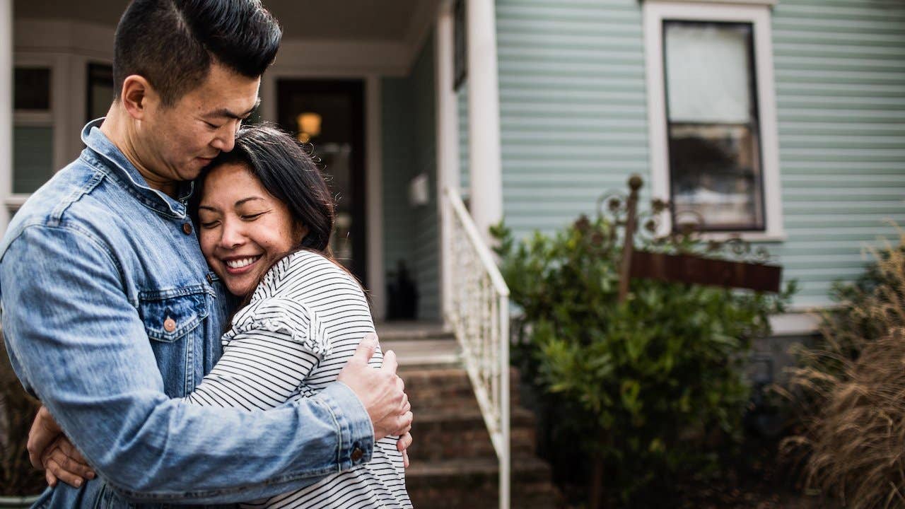 Couple hugging outside of light blue family home with a white railing leading up the front steps