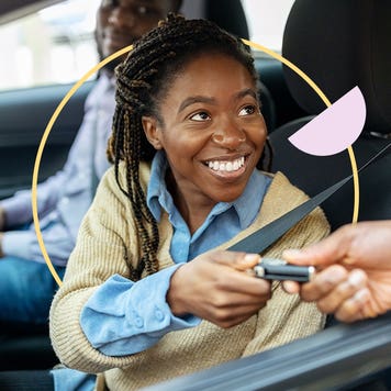 A young Black woman smiling as she takes a key while sitting in the driver's seat of a vehicle.