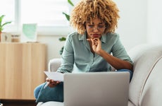 Serious Afro-American woman is sitting on a sofa and looking at a laptop while holding a paper in her hand. She is wearing glasses. She might be working from home or studying, or paying a utility bill. She is wearing a green shirt and jeans.