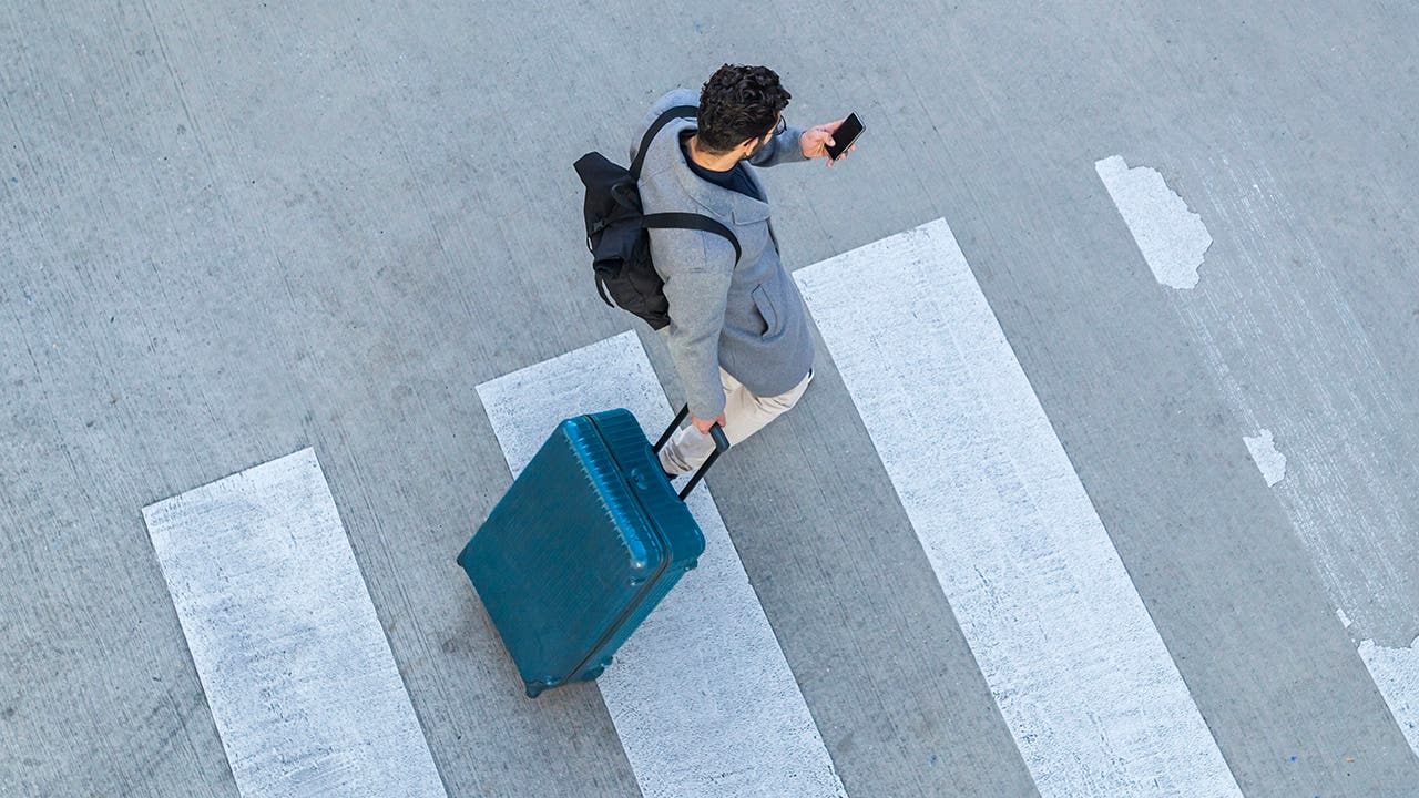 Businessman with baggage crossing the street while looking at cell phone, top view