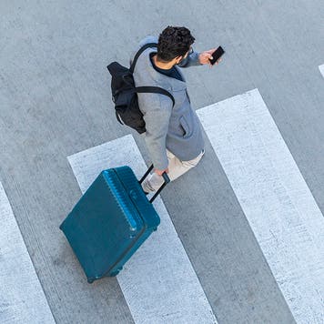 Businessman with baggage crossing the street while looking at cell phone, top view