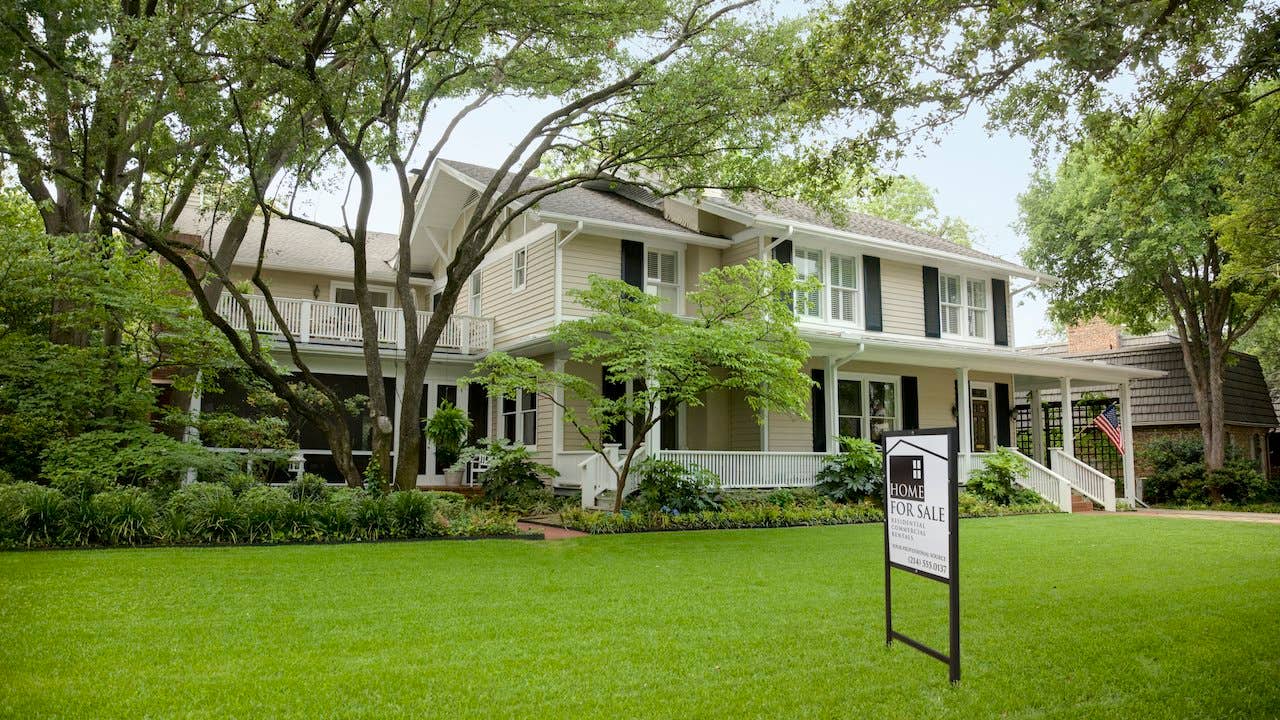 Exterior of large suburban home with for sale sign out front, with big green lawn and trees