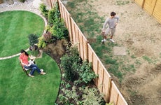 Woman sitting on one side of a yard while a man with a shovel stands, divided by a fence