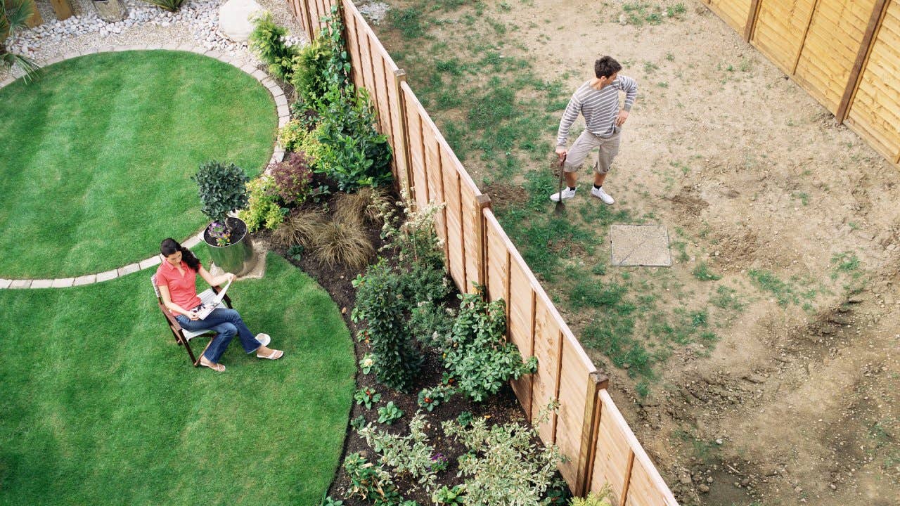 Woman sitting on one side of a yard while a man with a shovel stands, divided by a fence