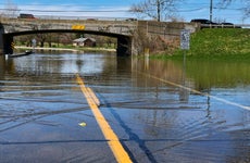 A flooded street leading under an overpass where a lot of the water is gathering. It is a bright and sunny day in what appears to be a small, rural area.