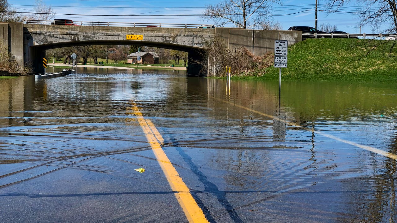 A flooded street leading under an overpass where a lot of the water is gathering. It is a bright and sunny day in what appears to be a small, rural area.