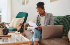 an african american woman reading papers sitting on her couch with her laptop