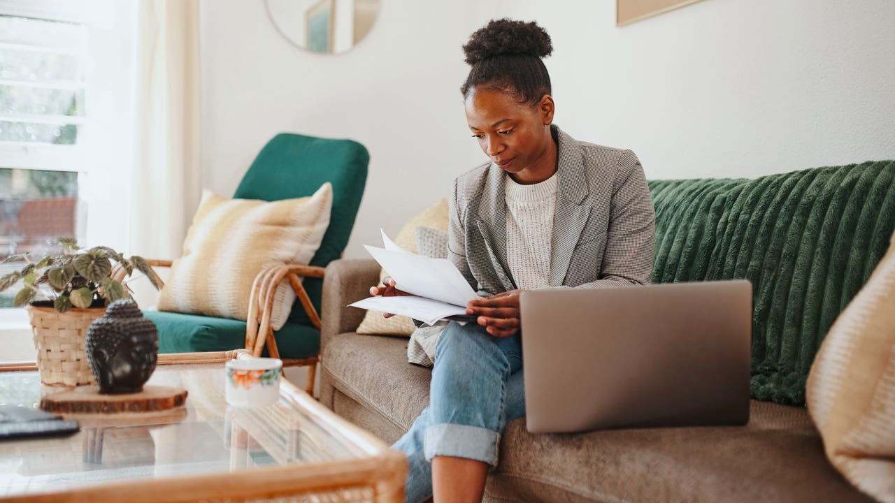 an african american woman reading papers sitting on her couch with her laptop