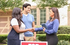 A young mixed-race couple shakes hands with a female real estate agent in front of a home that's for sale
