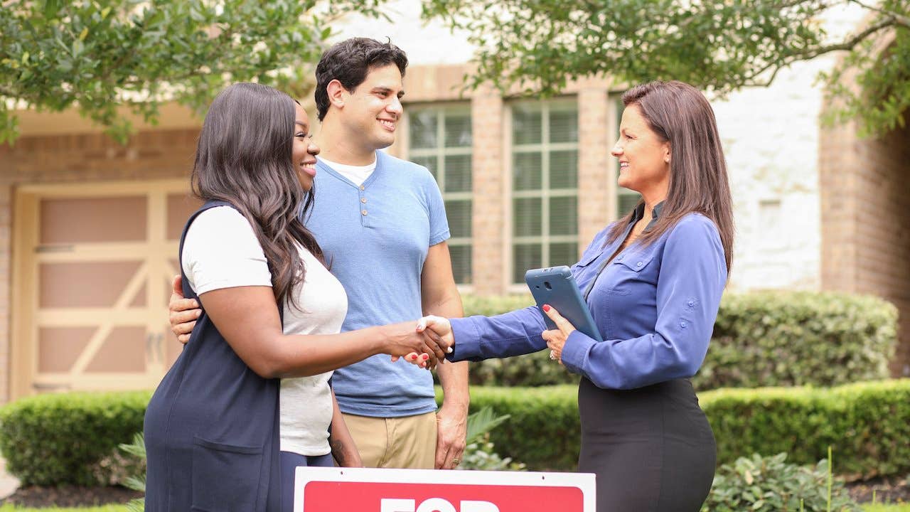 A young mixed-race couple shakes hands with a female real estate agent in front of a home that's for sale